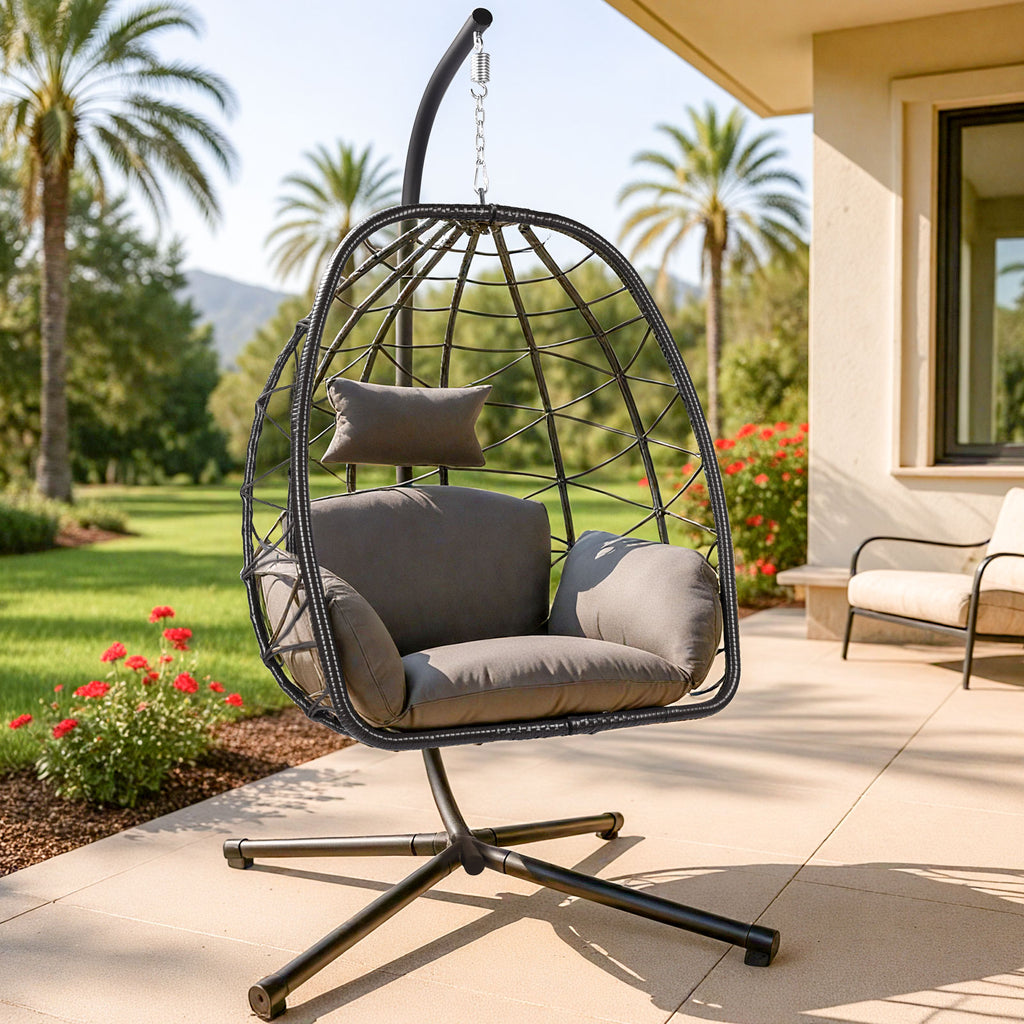 Hanging egg chair with gray cushions on a patio with palm trees and flowers in the background.