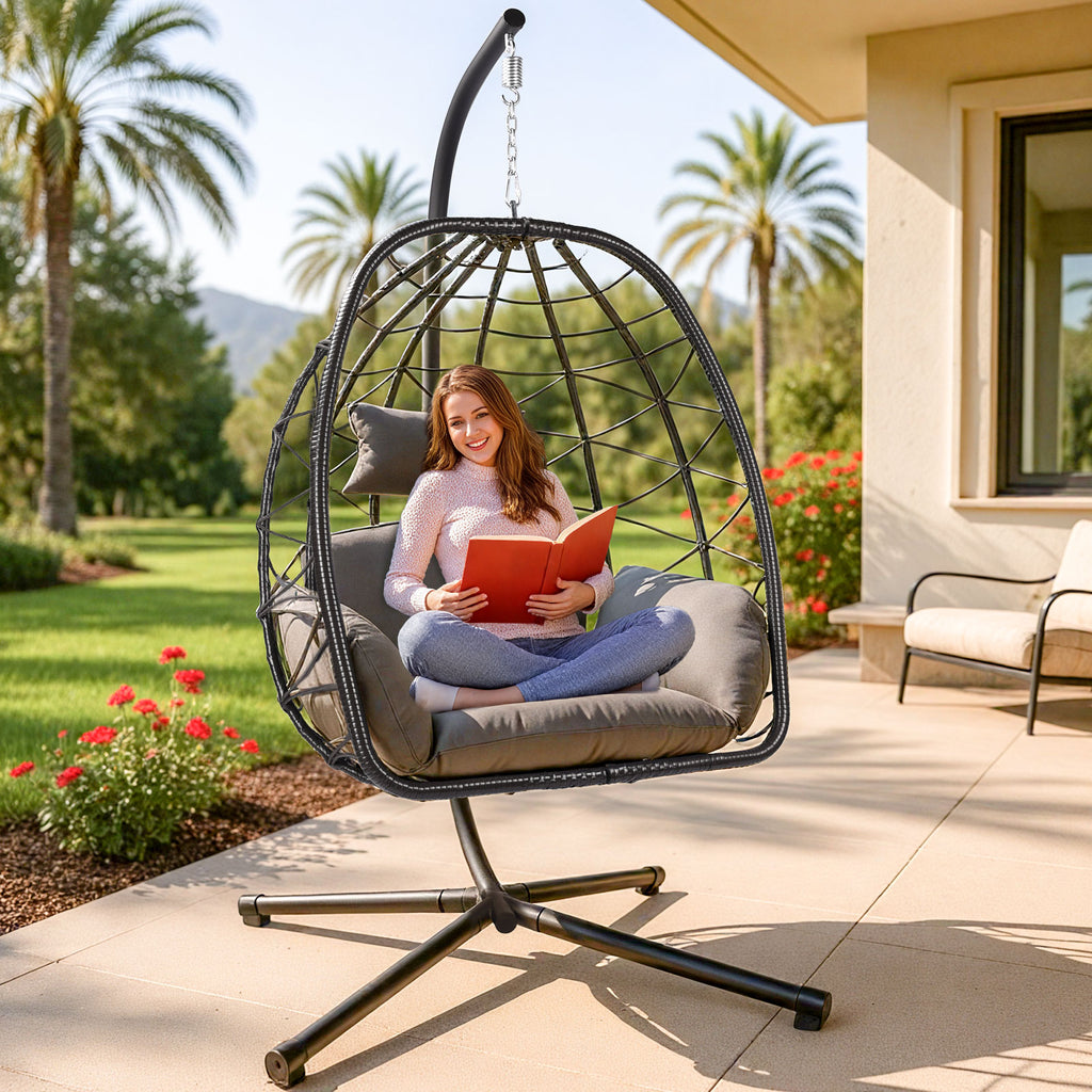 Person reading a book in a hanging egg-shaped chair on a patio with palm trees and flowers in the background.